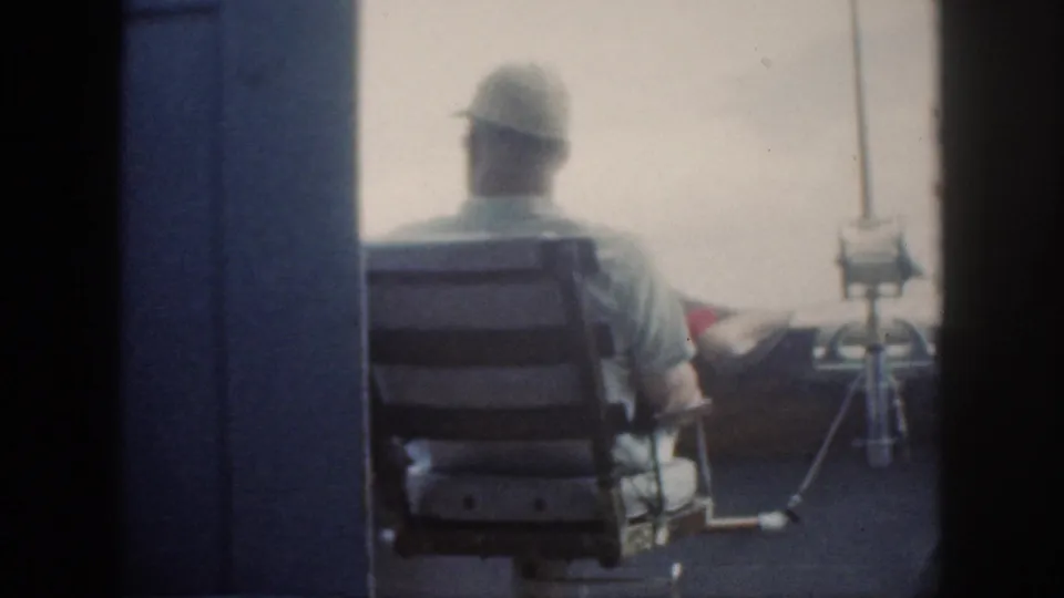 1959 Oahu boating frame with a cap-wearing adult in a deck chair, fishing rods upright, and pale sea in the background.