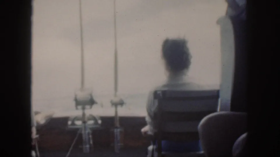 Faded 8mm frame from the 1959 Oahu clip showing a seated adult from behind, fishing gear, and bright water beyond the boat.