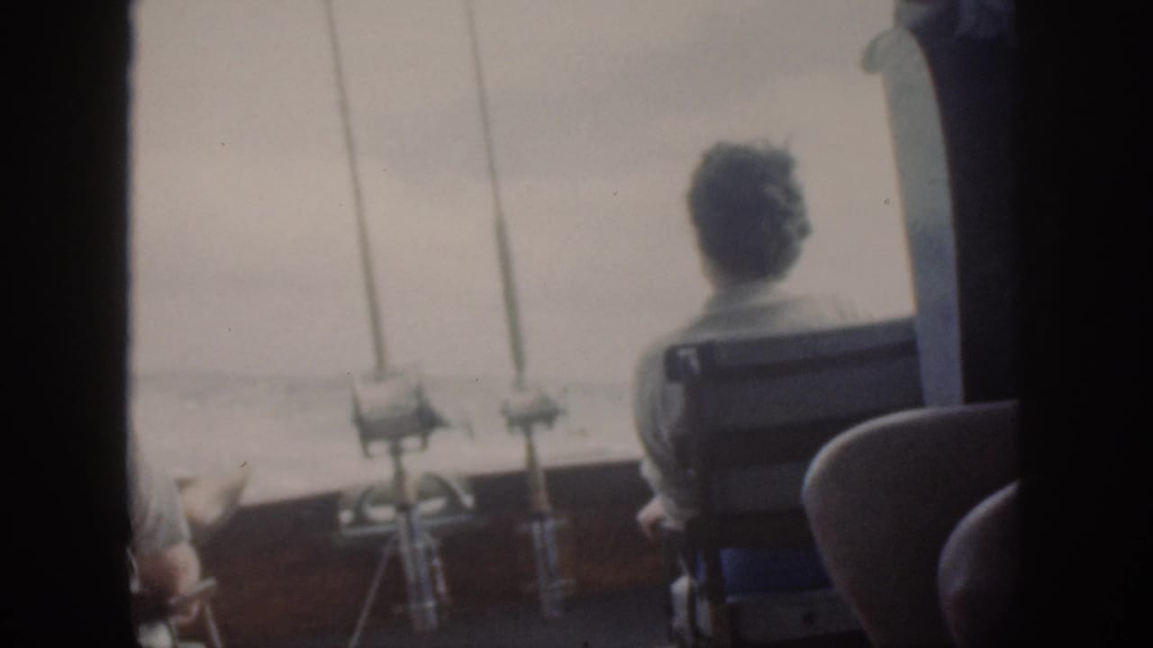 1959 Oahu home-movie frame showing passengers from behind on a boat, with mounted fishing rods and open water beyond.