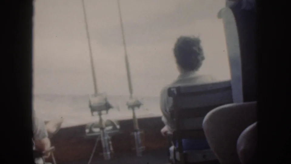 1959 Oahu home-movie frame showing passengers from behind on a boat, with mounted fishing rods and open water beyond.