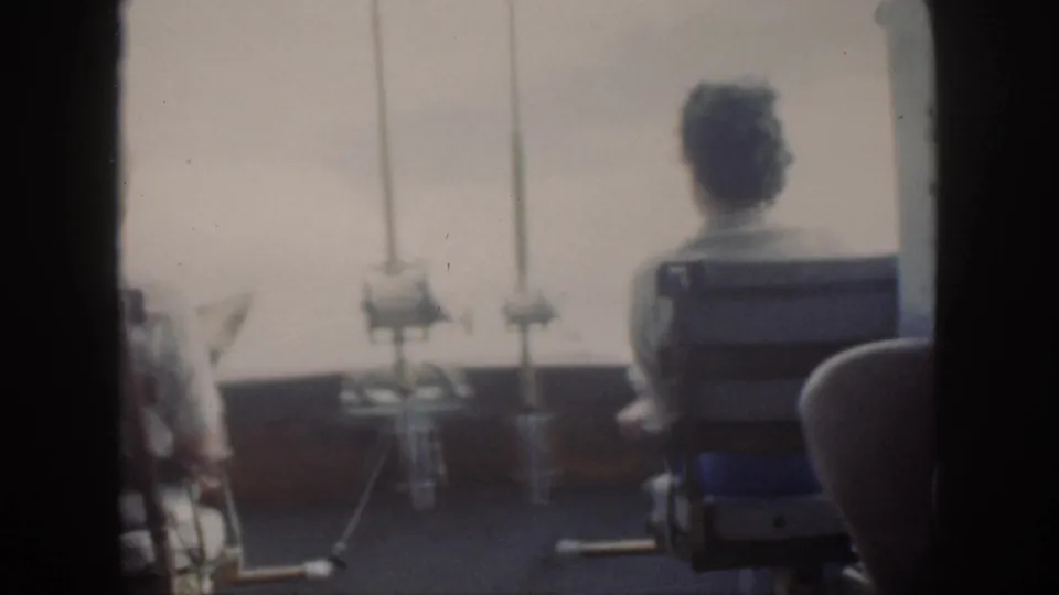 1959 Hawaii home-movie frame with a passenger seated on a boat, fishing gear mounted forward, and the sea in soft focus.