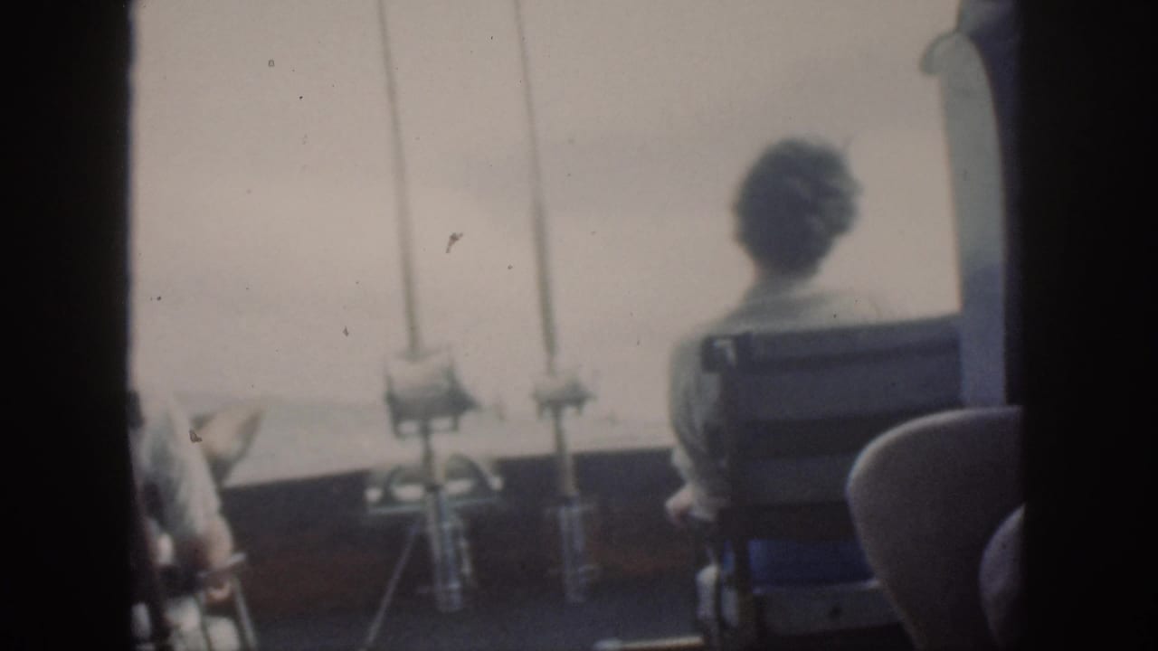 1959 Oahu home-movie frame showing seated adults from behind on a boat deck, facing bright water beside mounted fishing rods.