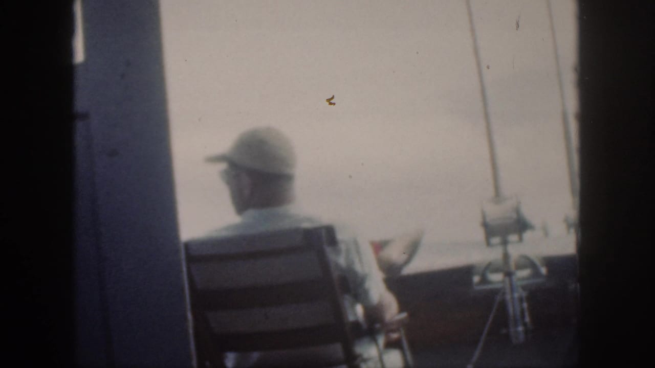 A man in a cap sits on a boat deck facing the water beside mounted fishing rods.