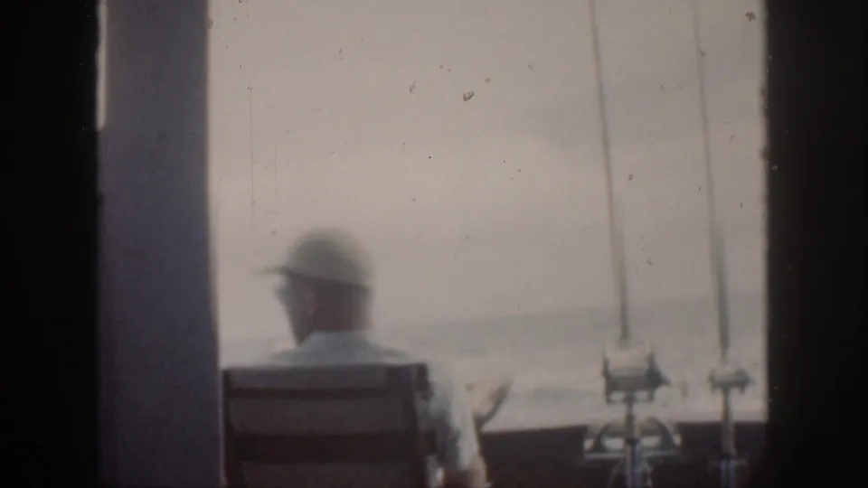 Soft 8mm frame from Oahu in 1959 showing a man in a cap seated near mounted rods, seen from behind against bright water.
