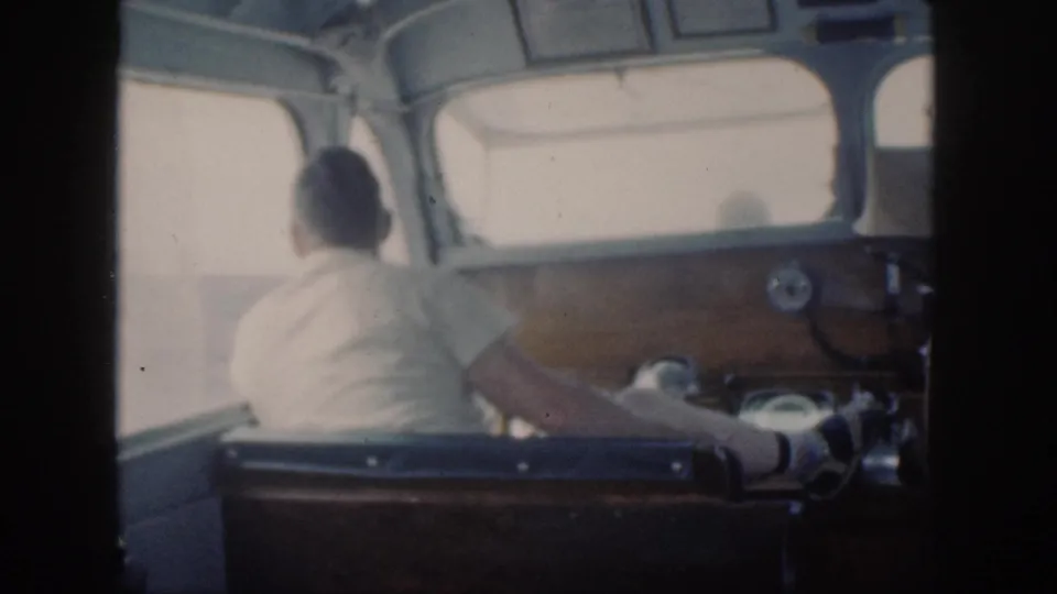 1959 Oahu home-movie frame showing a person at the wheel inside a small boat cabin, with windshield framing bright water outside.