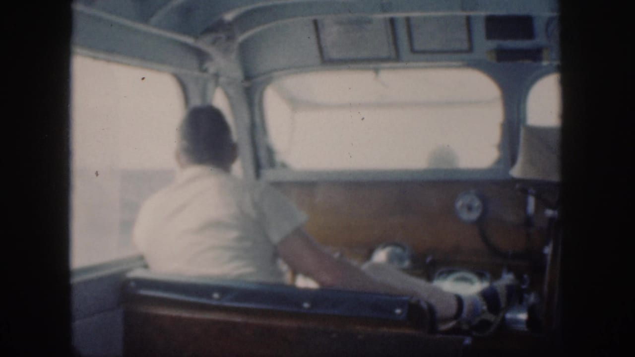 In 1959 Oahu home-movie footage, a man in a light shirt sits at the helm inside a small boat cabin.