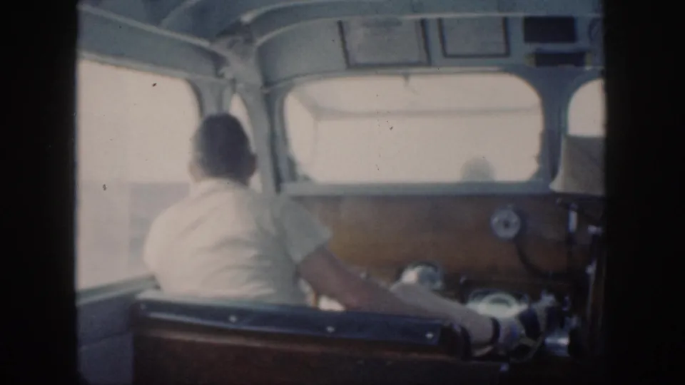In 1959 Oahu home-movie footage, a man in a light shirt sits at the helm inside a small boat cabin.