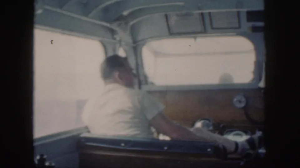 A light-shirted man sits forward at the boat controls, with the cockpit and bright seascape visible ahead.