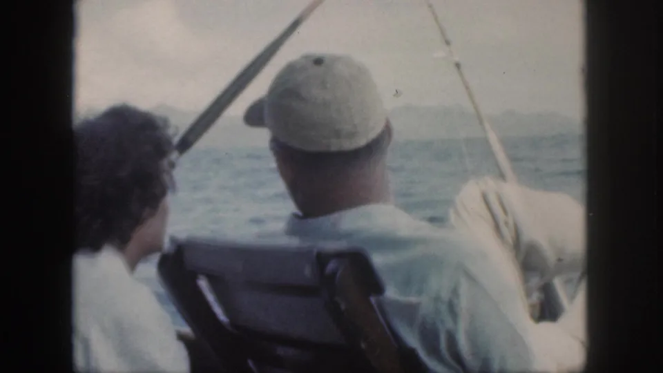 1959 Oahu home-movie still with a man in a light cap and a woman in white seated on a boat looking across the ocean.
