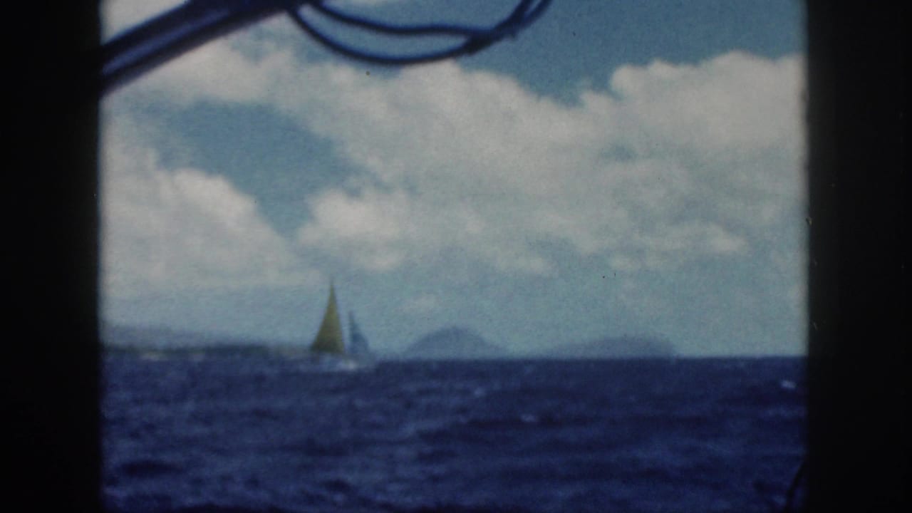 1959 Oahu 8mm frame of a small sailboat on open blue water, with distant island silhouettes and clouds seen from a boat.