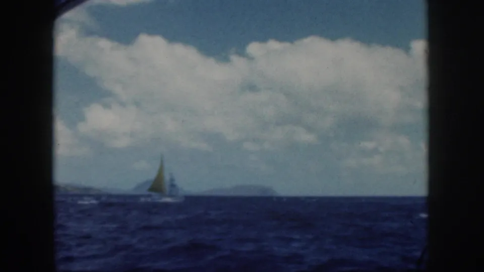 1959 Oahu home-movie frame showing a distant sailboat on blue water under a partly cloudy sky, with dark boat framing at the sides.