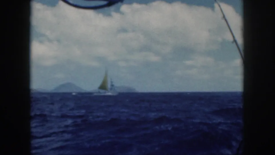 1959 Oahu 8mm view of a yellow-sailed boat crossing choppy ocean water, with a distant island shape under cloud cover.