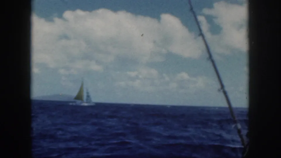1959 Oahu home-movie frame with a small sailboat near the horizon, cloudy sky, dark boat framing, and a fishing rod along the right side.
