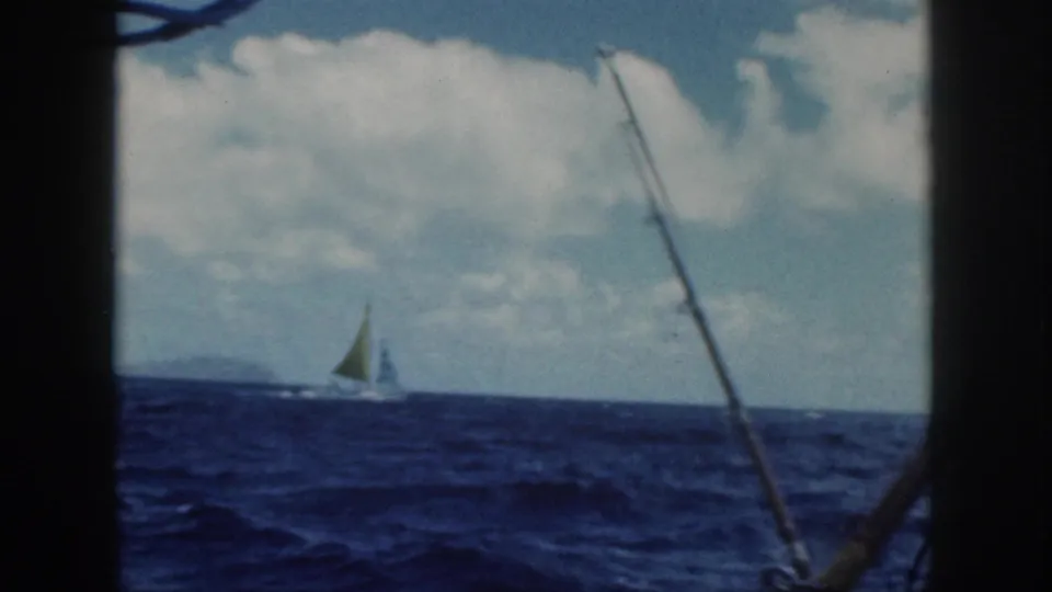 1959 Oahu boat view of a distant sailboat with a green sail crossing blue water under a cloudy sky.