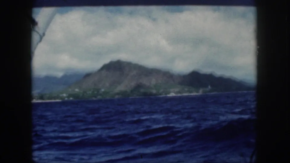 Offshore 1959 home-movie view of Oahu, with dark framing, rolling ocean, and volcanic mountains beneath cloud cover.
