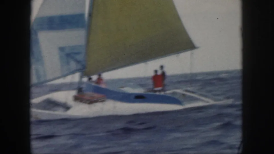1959 Oahu 8mm frame showing people aboard a white catamaran with yellow and blue sails on open water.