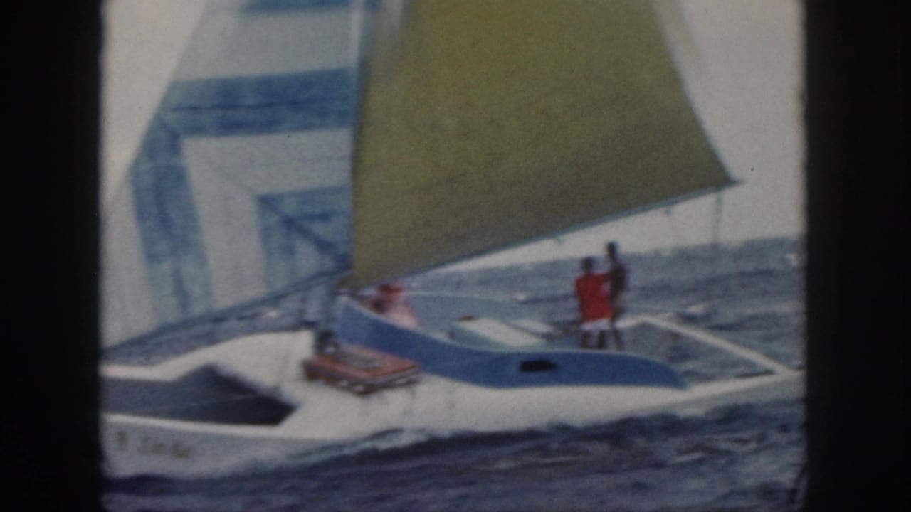 1959 Oahu home-movie frame of a small white sailboat with blue trim, yellow sail, and two people aboard on choppy water.