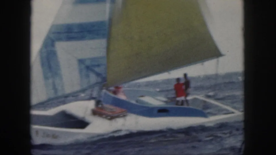1959 Oahu home-movie frame of a small white sailboat with blue trim, yellow sail, and two people aboard on choppy water.
