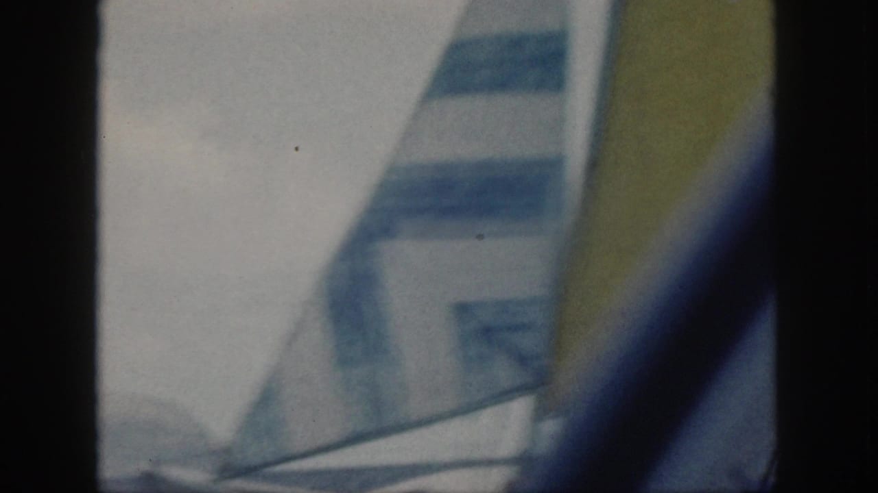 Soft 1959 Oahu 8mm frame with a striped sail and yellow sail edge crossing the view above ocean water.