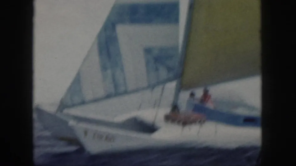 1959 Oahu home-movie side view of the small white sailboat under yellow and striped sails, with people visible near the deck.