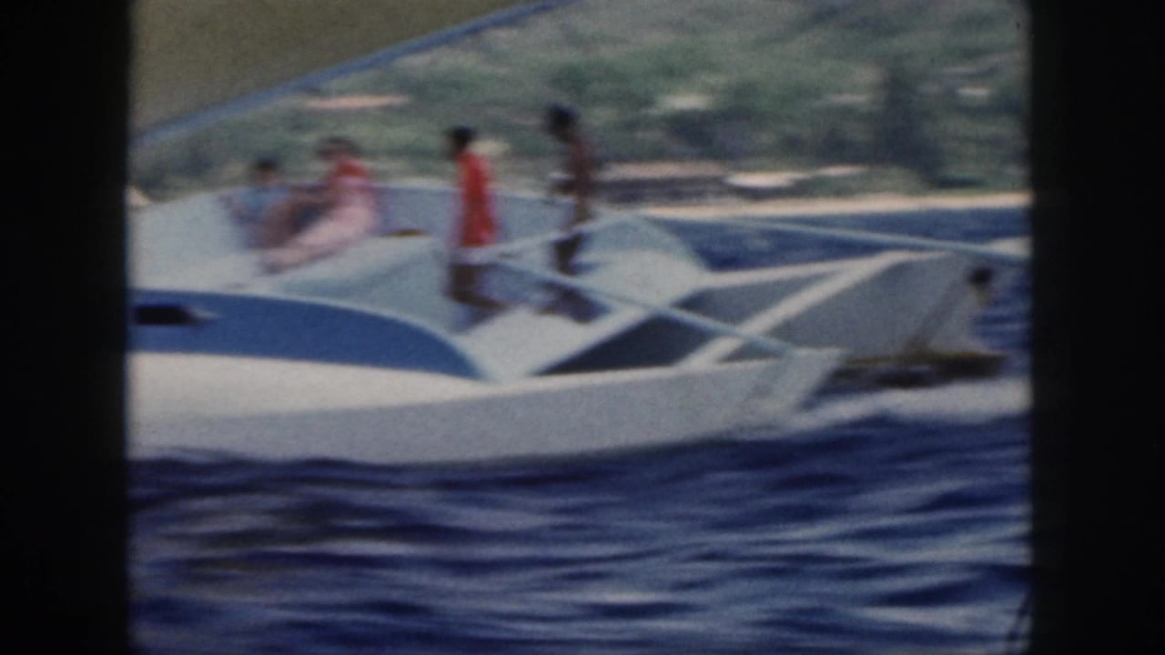 1959 Oahu home-movie frame showing a white-and-blue sailboat with people aboard crossing choppy water near a green shoreline.