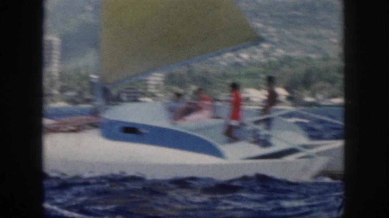 1959 Oahu frame of a white-and-blue sailboat under a yellow sail, with people on deck and shoreline buildings in the background.