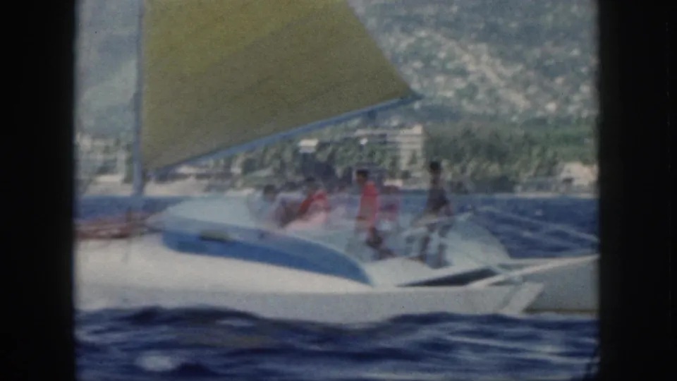 1959 Oahu 8mm frame showing a slightly blurred white-and-blue sailboat with a yellow sail and several people aboard.