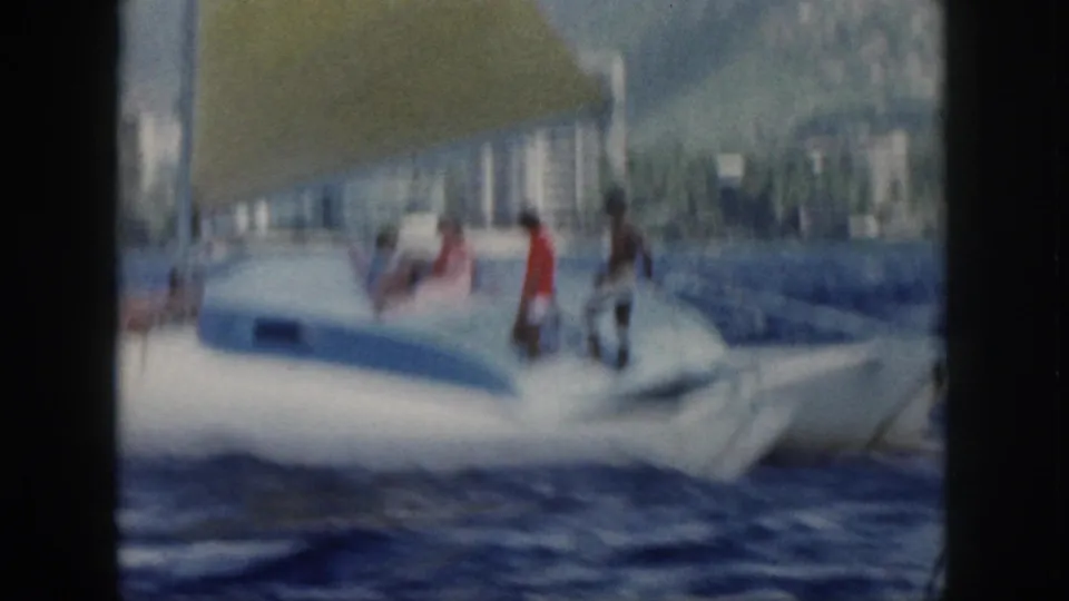 1959 Oahu home-movie frame of a sailboat making wake on choppy water, with people aboard beneath a yellow sail.