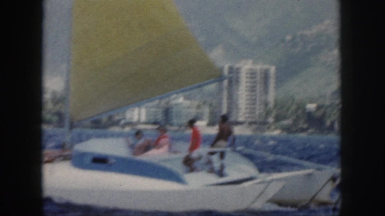 White sailboat with a yellow sail on blue water near a shoreline with tall buildings.