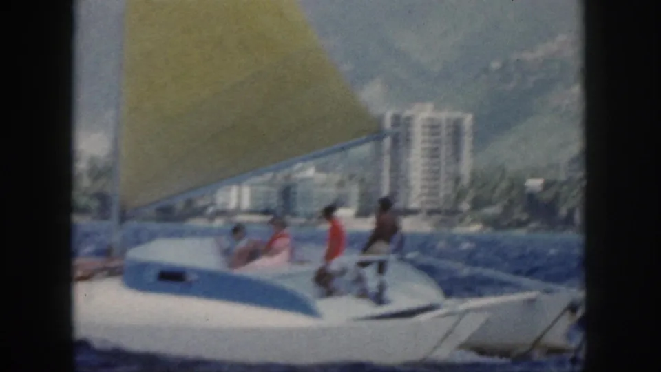 1959 Oahu home-movie frame showing a white recreational sailboat with a yellow sail and passengers offshore from a developed shoreline.
