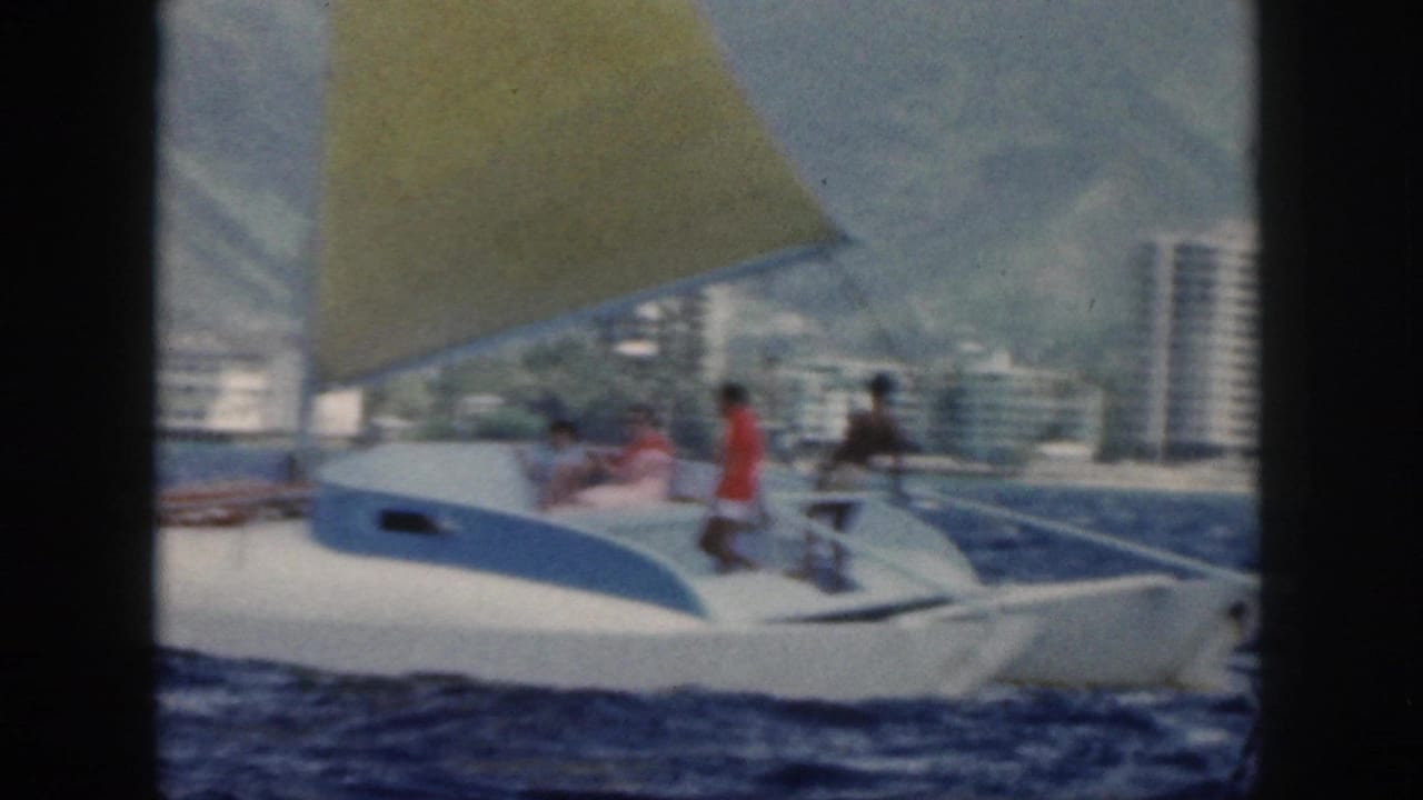Small sailboat carrying several people, with high-rise buildings and mountains behind it.