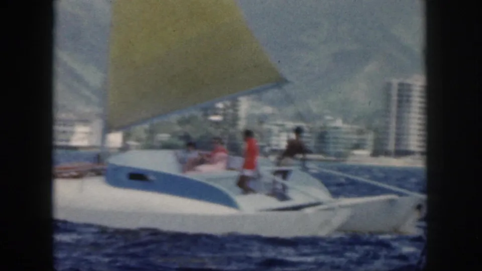 White sailboat with several visible passengers gliding offshore in Oahu, with high-rise buildings and mountains in the background.