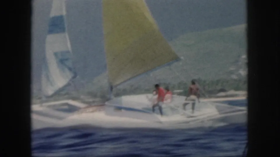 Soft 8mm frame of the yellow-sailed boat moving across choppy water off Oahu, with passengers seated on the deck.