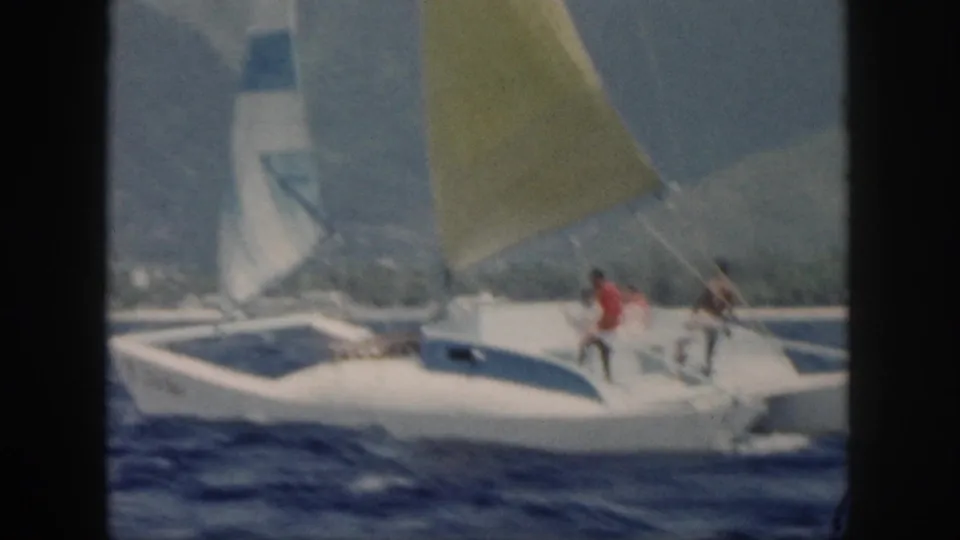 Oahu 1959 sailing frame showing a white boat, yellow sail, visible passengers, blue sea, and hazy coastal mountains.