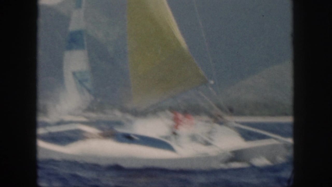 1959 Oahu home-movie frame of a white catamaran with blue trim and yellow sail crossing choppy ocean water.