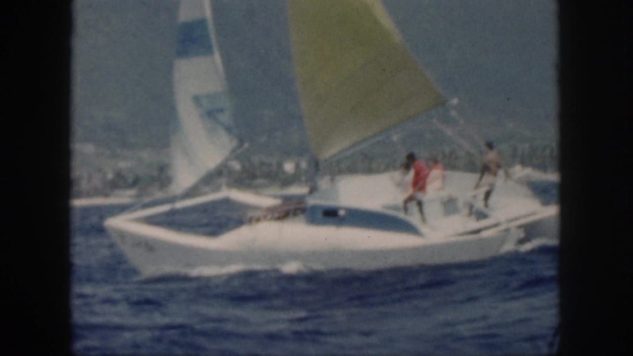 Catamaran in profile with several people on deck and sails raised over dark blue water.