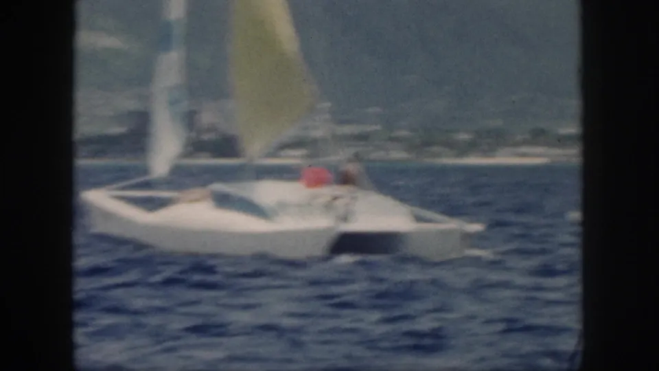1959 Oahu frame of the catamaran farther away on blue water, with its yellow sail and coastline in the background.