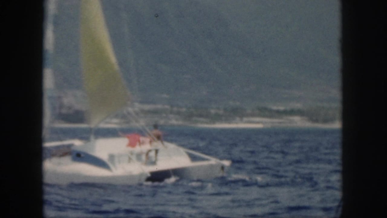 1959 Oahu home-movie frame showing a white catamaran with a yellow sail and two distant people aboard on rough blue water.