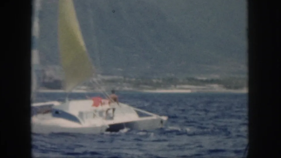 1959 Oahu home-movie frame showing a white catamaran with a yellow sail and two distant people aboard on rough blue water.