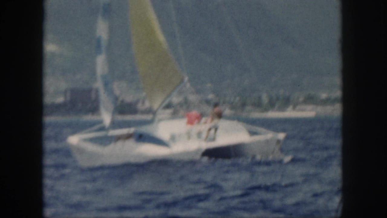 Catamaran with two people aboard crossing choppy water under hazy sky.