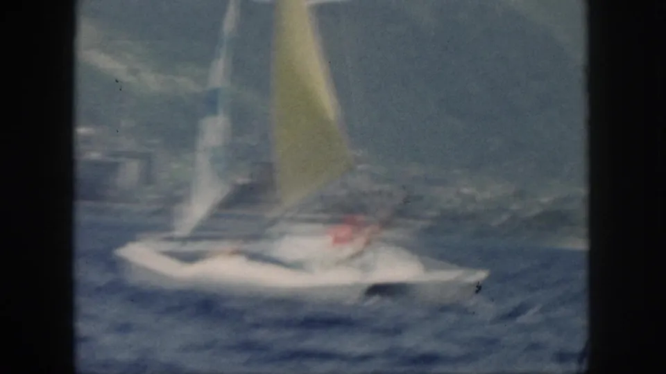 Blurred 1959 Oahu home-movie frame with a white catamaran and yellow sail moving quickly across choppy blue water.