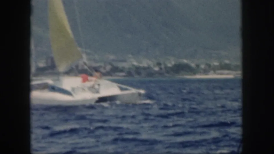 1959 Oahu frame showing the catamaran angled near the distant shoreline, with two people aboard and wind-ruffled water in front.
