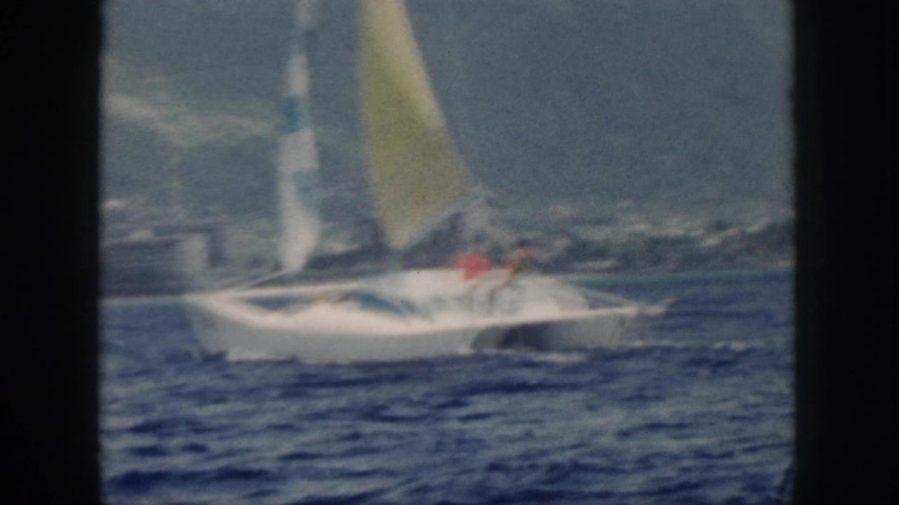 1959 Oahu home-movie frame showing a white catamaran with a yellow sail moving across blue water near a mountainous shoreline.