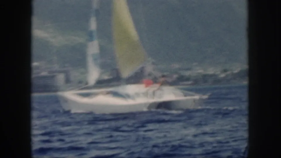 1959 Oahu home-movie frame showing a white catamaran with a yellow sail moving across blue water near a mountainous shoreline.