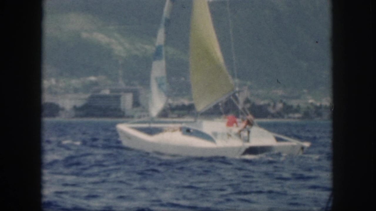 Catamaran with two people aboard sailing near coastal buildings and mountains.