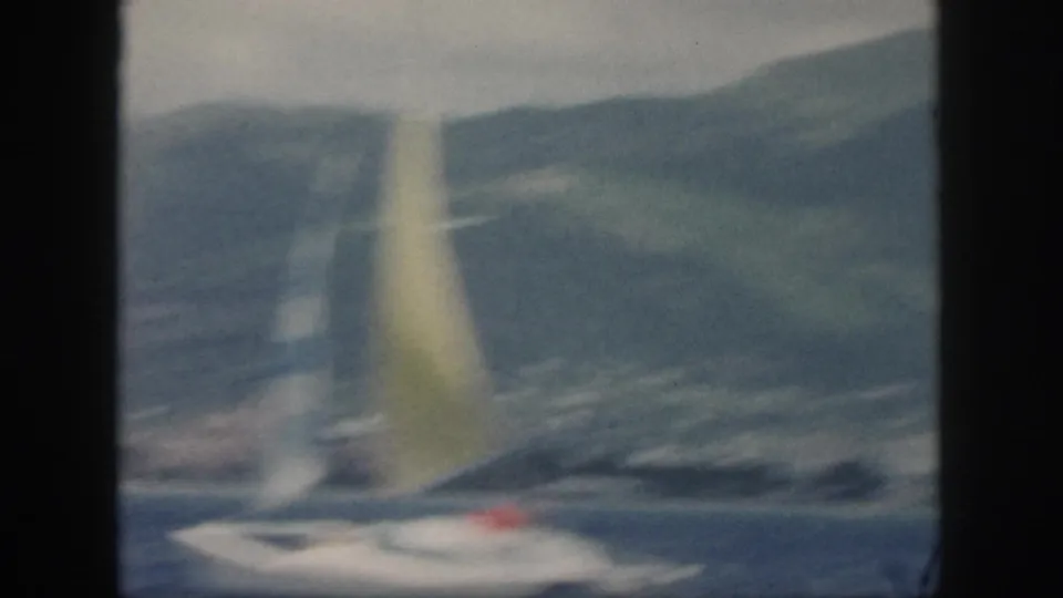 Blurred 1959 Oahu home-movie frame of a catamaran under a yellow sail, seen against blue water and a mountainous coast.