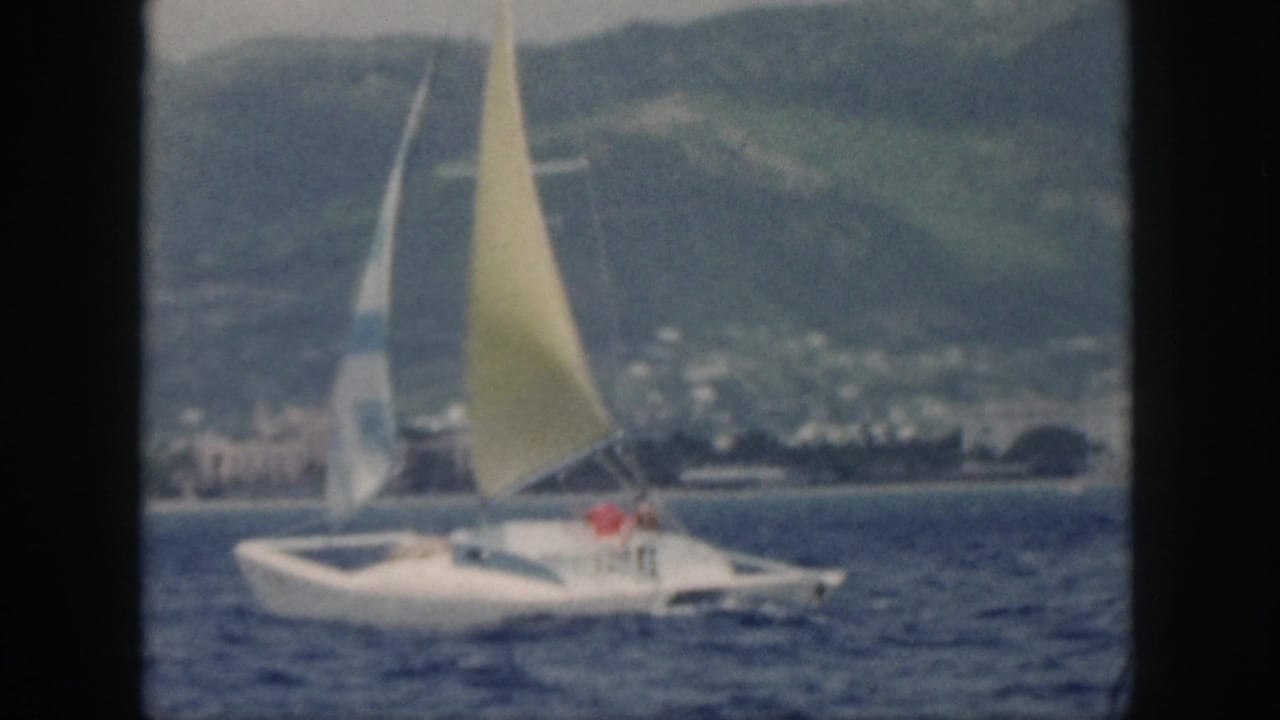 1959 Oahu home-movie frame showing a white sailboat with a yellow mainsail and striped jib offshore from a green coastline.