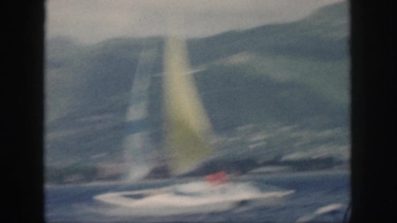 Soft 1959 Oahu frame of the sailboat passing across the water, with the yellow sail and coastal hills visible through motion blur.