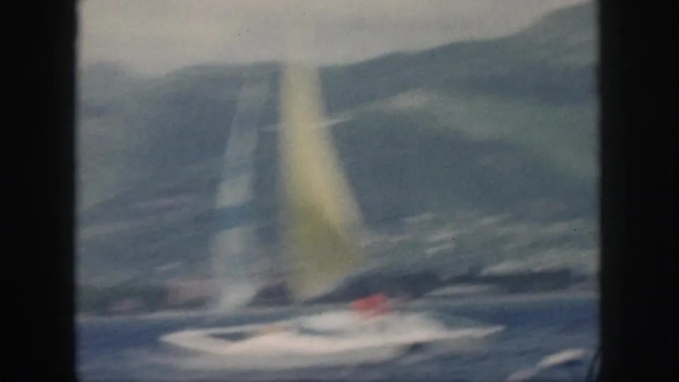 Soft 1959 Oahu frame of the sailboat passing across the water, with the yellow sail and coastal hills visible through motion blur.
