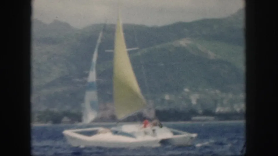 1959 Oahu frame showing the white sailboat under a yellow mainsail, with a hazy mountainous shoreline and coastal settlement behind it.